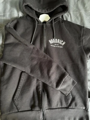 Svart zip hoodie från Hoodrich - Snygg svart hoodie med dragkedja från Hoodrich. Tryck med texten 'HOODRICH EST. 2014 WELCOME TO THE FAMILY' både fram och bak. Utrustad med huva och snören samt två fickor framtill. Tillverkad i mjukt bomullsmaterial för skön känsla.