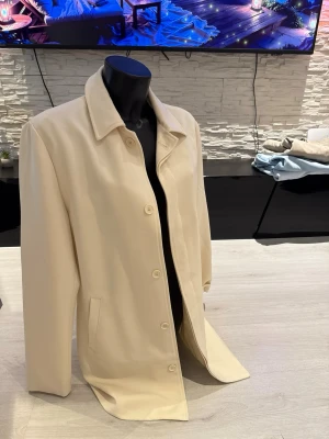 Beige klassisk rock med knappar - Stilren beige rock med rak passform och klassisk krage. Rocken har knäppning framtill med fem knappar och två sidofickor. Materialet ser ut att vara mjukt och slätt, perfekt för en clean och tidlös look.