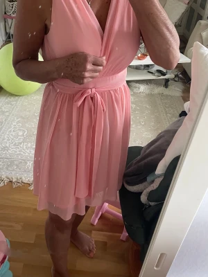 Rosa omlottklänning med knytband - Chiffongklänning från Zoul edition omlottdesign framtill och knytband i midjan. Klänningen har en V-ringning och är ärmlös. Baktill finns en dragkedja och en öppning i ryggen.färgen går mer åt laxrosa .har haft på mig två gånger, klänningen är så fin skick 