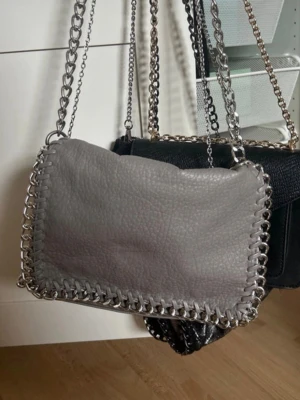 Grå handväska med kedjerem - Snygg grå handväska i mjukt skinnliknande material med silverfärgade kedjedetaljer runt kanterna och som axelrem. Väskan har en rektangulär form och ger en trendig och cool vibe. Perfekt accessoar för att lyfta din outfit.