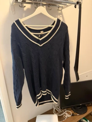 Mörkblå stickad tröja Divided H&M - Mörkblå stickad tröja från Divided H&M med vita ränder vid v-ringad hals, ärmslut och nederkant. Tröjan har ett klassiskt kabelstickat mönster och är långärmad. Perfekt för en avslappnad och trendig look.