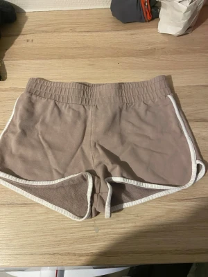 Beige mjukisshorts med vit kant - Säljer ett par beige mjukisshorts med vit kant längs sidorna och nederkanten. Shortsen har elastisk midja och är i ett mjukt material som passar perfekt för chill dagar eller träning.