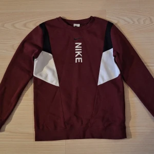 Vinröd Nike sweatshirt med logga stl 158-170 - Nyskick, snvänd ett fåtal gånger. Snygg vinröd sweatshirt från Nike med vit logga framtill. Tröjan har svarta och vita paneler på axlar och sidor, rund hals och långa ärmar. Perfekt för en sportig och avslappnad stil.