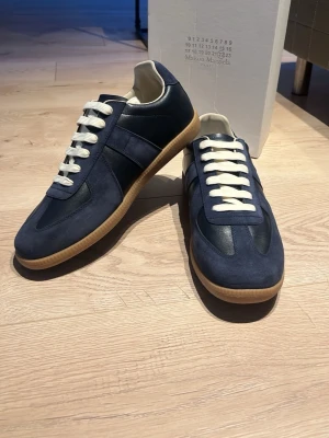 Maison Margiela - Snygga blå sneakers från Maison Margiela med ovandel i mocka och läder, vita snören och klassisk gummisula. Skorna har en stilren design med diskreta detaljer och passar perfekt till jeans eller chinos.