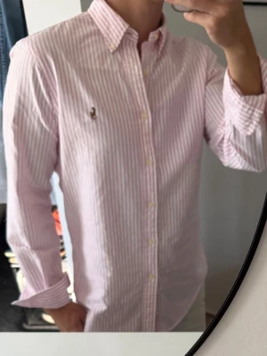 Rosa och vit randig skjorta - Säljer en klassisk rosa och vit randig skjorta med button-down krage och långa ärmar. Skjortan har knappar framtill och en liten broderad logga på bröstet. Perfekt för en clean och stilren look. Väldigt missfärgad i armhålorna som man ser på sista bilderna, därav priset 