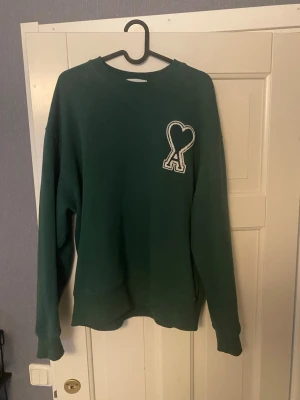 Grön AMI PARIS sweatshirt - Grön sweatshirt från Ami Paris med det klassiska hjärta-A broderiet på bröstet. Enkel och stilren design som är lätt att matcha. Säljer då den är för liten på mig. Priset ej hugget i sten. Tveka inte på att skriva om du har funderingar 