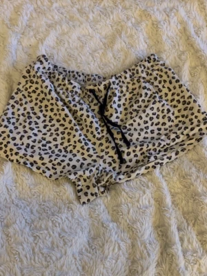 Leopardmönstrade pyjamasshorts - Säljer ett par pyjamasshorts från NAP QUEEN i leopardmönster. Shortsen är vita med svarta och bruna fläckar, har resår i midjan och svart dragsko för justerbar passform. Perfekta för mysiga hemmakvällar eller lata morgnar.
