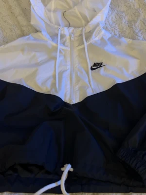 Vit och marinblå vindjacka från Nike - Snygg vindjacka från Nike i vitt och svart med halv dragkedja framtill och justerbar huva. Jackan har elastiska muddar och dragsko i nederkant för skön passform. Perfekt för blåsiga dagar och sportiga outfits.