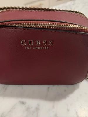 Vinröd axelväska från Guess - Snygg vinröd axelväska från Guess med guldfärgade detaljer och kedjerem. Väskan har två fack med dragkedja och Guess-logga framtill. Materialet är strukturerat konstläder och remmen har en bekväm axelplatta i matchande färg.