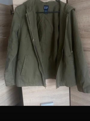 Olivgrön höstjacka från GAP - Snygg olivgrön jacka från GAP med huva och dragsko. Jackan har dragkedja framtill, två snedställda fickor och raka ärmar. Perfekt nu till hösten 