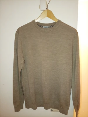 J. Lindebergh Merino  - En ny oanvänd stilren bekväm beige stickad tröja med rund hals och långa ärmar. Tröjan har en enkel design utan mönster och är tillverkad i en mjuk ullmix som passar perfekt till lager på lager storlek m märke av J.Lindeberg butikspris 1399 