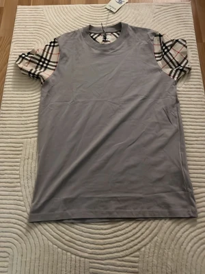 Grå t-shirt med rutiga ärmar Burberry - Snygg grå t-shirt från Burberry med klassiska rutiga korta ärmar i beige, svart och rött. Perfekt för dig som vill ha en stilren men ändå unik look. Tillverkad i mjuk bomull för skön känsla hela dagen.