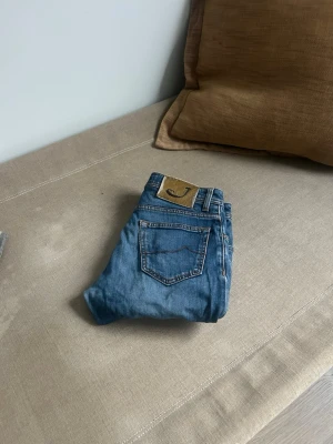 Blå raka jeans med läderpatch - Säljer ett par klassiska blå jeans i rak modell med fem fickor och snygga slitningar. Jeansen har en läderpatch bak i midjan och kontrastsömmar. Perfekta till vardags för en avslappnad look. Materialet är denim i bomull.