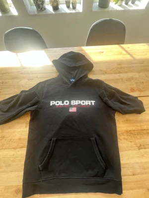 Svart hoodie från Polo Sport - Säljer en svart hoodie från Polo Sport Ralph Lauren med stor ficka framtill och huva. Tryck med logga och amerikansk flagga på bröstet. Tillverkad i mjuk bomull, perfekt för en avslappnad och sportig stil.