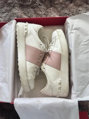 Rosa valentino sneakers  - Snygga vita sneakers från Valentino med ett brett ljusrosa band över mitten och coola nitar på sulan. Skorna har vita snören och en chunky sula som ger en modern vibe. Perfekta för dig som vill ha något stilrent men ändå unikt. Pris kan absolut diskuteras vid snabb affär