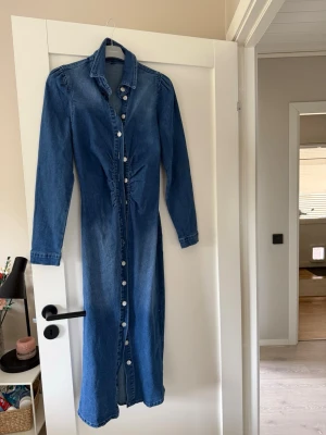 Lång jeansklänning med knappar - Säljer en lång jeansklänning i blå denim med långa ärmar. Klänningen är inte använd och tyvärr vet jag inte vart den är ifrån eller vilken storlek. Skulle säga mellan S och M. 