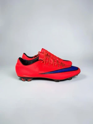 Röda fotbollsskor med blå detalj - Snygga röda fotbollsskor med blå Nike-logga på sidan och svarta detaljer vid hälen. Skorna har snörning och dobbar för spel på gräs. Tillverkade i syntetmaterial för bra grepp och lätt känsla på planen.