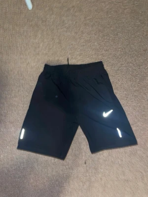 Svarta träningsshorts  - Säljer ett par svarta träningsshorts  med reflexdetaljer och snörning i midjan. Shortsen har en klassisk passform och är tillverkade i ett lätt och snabbtorkande material, perfekta för träning eller sport.