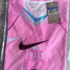 Rosa FC Barcelona matchtröja Nike - Snygg rosa FC Barcelona matchtröja från Nike med klubbmärke och sponsorlogga på bröstet. Tröjan har vita och ljusblå detaljer, V-ringning med blå kant och tunna vertikala ränder. Perfekt för fotbollsträning eller att bära som supporter.