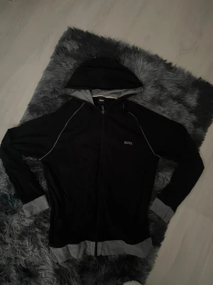 Svart hoodie med dragkedja från BOSS - Snygg svart hoodie från BOSS med dragkedja framtill och kontrasterande grå muddar och fåll. Hoodien har en huva med grå insida och diskret BOSS-logga på bröstet. Perfekt för en chill och stilren look.