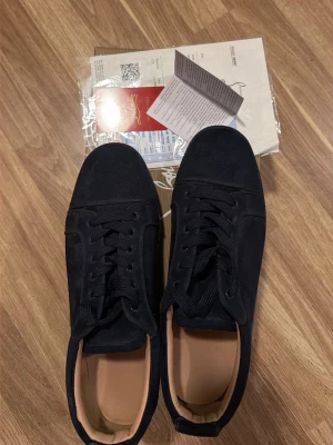 Svarta sneakers i mocka med röd sula - Stilrena svarta sneakers från Christian Louboutin i mjuk mocka med klassisk röd sula och svarta skosnören. Insidan är beige och designen är lågskuren med rund tå. Perfekta för dig som vill ha en lyxig och tidlös look.