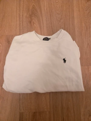Vit sweatshirt från Polo Ralph Lauren - Vit sweatshirt med rund hals och svart broderad logga på bröstet från Polo Ralph Lauren. Klassisk modell med ribbade muddar vid ärmslut och nederkant. Perfekt för en clean och stilren look.