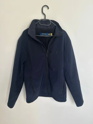 Mörkblå vindjacka från Polo Ralph Lauren - Stilren mörkblå vindjacka från Polo Ralph Lauren med hög krage och dragkedja framtill. Jackan har tre fickor med dragkedja och diskret logga på bröstet. Perfekt för blåsiga dagar och enkel att matcha till olika outfits.