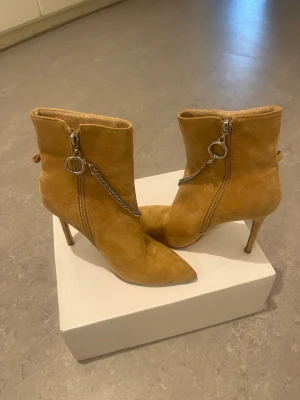 Camelbruna stövletter med kedjedetalj  - Snygga camelbruna stövletter i mocka med spetsig tå och smal klack. Dragkedja på sidan med silverfärgad ring och kedja som dekoration. Perfekta för dig som vill ha en edgy och trendig look. Använda ett par gånger nästan som ny😊 