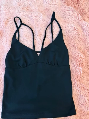 Svart crop top med tunna axelband - Snygg svart crop top med tunna, justerbara axelband och djup v-ringning både fram och bak. Toppen har en figurnära passform och är tillverkad i ett stretchigt material som känns bekvämt mot huden. Perfekt att matcha med jeans eller kjol.