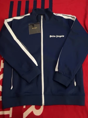 Blå ziptröja från Palm Angels - Snygg blå ziptröja från Palm Angels med vita detaljer längs ärmarna och vit dragkedja. Tröjan har hög krage, två fickor med dragkedja och logga på bröstet. Perfekt för en sportig och trendig look. Pris kan diskuteras och mer bilder kan skickas 