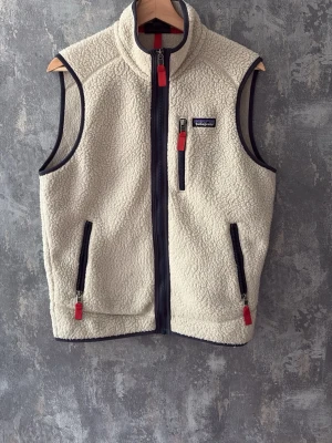 Patagonia väst  - Säljer en beige fleeceväst från Patagonia med svarta detaljer och röda dragkedjor. Västen har hög krage, tre fickor med dragkedja och mjukt teddy-material. Perfekt för lager-på-lager och chill stil, knappt använd då den inte passa, kvitto finns.