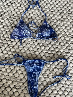 Blå bikini med paisleymönster och knyt - Säljer en blå bikini med vitt paisleymönster. Överdelen är trekantsmodell med knyt i nacken och ryggen, och underdelen har smala knytband i sidorna. Perfekt för sommaren och strandhäng! Helt nytt skick. 1 del = 65 kr, 2 delar = 110 kr
