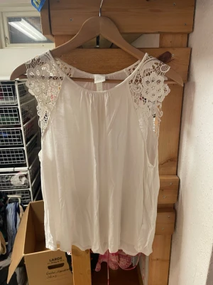 Vit topp med spetsdetaljer - Säljer en luftig vit topp med vackra virkade spetsdetaljer på axlarna. Toppen har rund halsringning och är ärmlös, vilket ger en fräsch och somrig look. Perfekt att matcha med jeans eller kjol för en trendig outfit.
