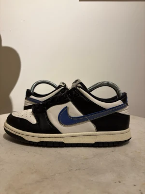 Nike Dunk Low svart/vit/blå - Snygga Nike Dunk Low sneakers i svart, vitt och blått. Skorna har klassisk låg siluett, svart läder på tå och häl, vit bas samt blå swoosh på sidan. Perforerad tåbox för ventilation och svart yttersula. Perfekta för dig som gillar streetwear och stilrena färger.