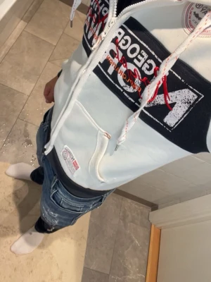 Vit hoodie Geographical Norway - Snygg vit hoodie med dragkedja från Geographical Norway. Stor norsk flagga och tryck på bröstet, samt broderade detaljer på ärmar och rygg. Marinblå muddar och innerfoder i huvan. Praktiska fickor framtill och justerbar huva med snören.