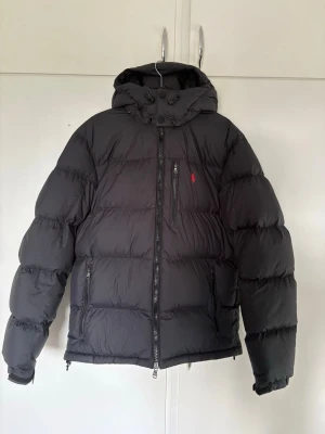 Vinterjacka från Ralph Lauren  - Säljer en svart pufferjacka från Ralph Lauren med huva och dragkedja framtill. Jackan har tre fickor med dragkedja och en liten röd logga på bröstet. Puffig design och elastiska muddar vid ärmsluten. Perfekt för kalla vinterdagar. Nypris 5000 på boozt