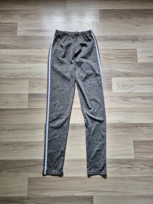 Grå träningsleggings med vita sidoränder - Snygga grå träningsleggings med vita ränder längs sidorna och elastisk midja. Perfekta för en sportig och avslappnad stil. Mjukt och stretchigt material som sitter skönt hela dagen.