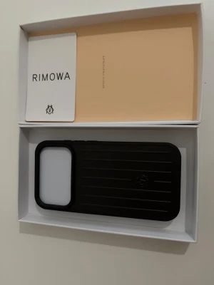 Svart mobilskal från Rimowa iphone 17 PRO - Snyggt svart mobilskal från Rimowa med räfflad design och diskret logga på baksidan. Skalet är tillverkat i ett tåligt material och har en rektangulär form med stor öppning för kameran. Perfekt för dig som vill ha en stilren och exklusiv look på mobilen.