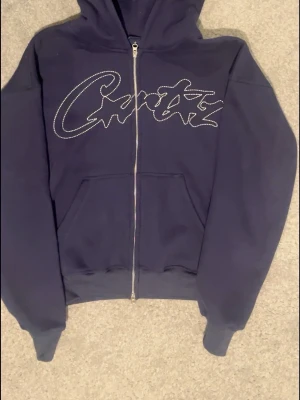 Mörkblå Corteiz zip hoodie - Mörkblå hoodie med dragkedja och stor ficka framtill. Snyggt tryck i vitt på bröstet i form av ett grafiskt mönster. Mjuk insida och huva för extra komfort. Perfekt för en avslappnad streetwear-look.