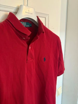 Röd pikétröja från Polo Ralph Lauren - Klassisk röd pikétröja från Polo Ralph Lauren med blå häst broderad logga på bröstet. Tröjan har korta ärmar, krage och två knappar vid halsen. 