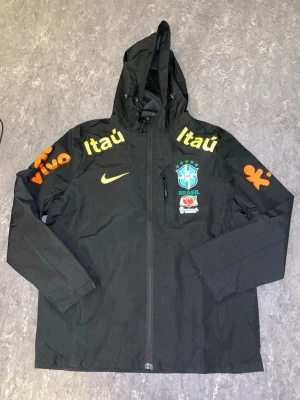 Svart Brasil Nike vindjacka med huva - Snygg svart vindjacka från Nike med Brasil-logga och sponsormärken i gult och orange. Jackan har huva, dragkedja framtill och tryck på både bröst och ärmar. Perfekt för dig som gillar fotboll och streetwear.