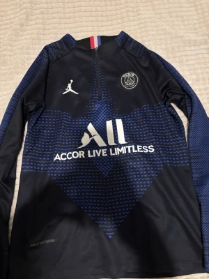PSG långärmad träningströja blå - Snygg långärmad träningströja från PSG i mörkblått med blått mönster och vita loggor. Tröjan har dragkedja vid halsen, PSG-emblem och Jordan-logga på bröstet samt texten 'Accor Live Limitless' framtill. Tillverkad i funktionsmaterial.
