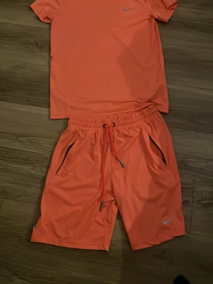 Nike träningsset i orange - Säljer ett sportigt träningsset från Nike i stark orange färg. Setet består av en t-shirt och shorts i lätt och snabbtorkande material med Dri-FIT-teknologi. Shortsen har dragsko i midjan och praktiska fickor. Perfekt för gym, löpning eller andra sportaktiviteter.