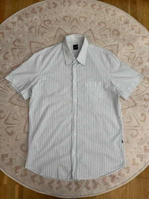 Hugo Boss skjorta  - Storlek: L Färg: Vit/ljusblå Skick: Mycket bra  Klassisk skjorta från BOSS med stilrent blåvitt randigt mönster och kort ärm. Slim fit-modell med bröstficka och diskret BOSS-detalj på sidan. Enkel och stilren modell som passar till både vardag och finare tillfällen.  Se bilder för skick och detaljer.