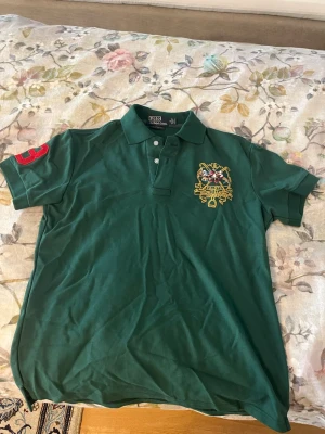 Grön pikétröja polo - Snygg grön pikétröja med broderat emblem på bröstet och polospelare på ryggen. Tröjan har krage, knappar framtill och korta ärmar. Detaljer i rött på ena ärmen och broderade inslag i flera färger. Den behöver stryckjärn sedan är den som ny, legat i mitt skåp ganska länge.