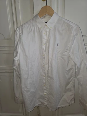 Vit skjorta från Gant - Klassisk vit skjorta från Gant storlek 170, dvs. för kille i tonåren, med button down-krage och broderad logga på bröstet. Skjortan har långa ärmar och knäppning framtill. Perfekt för en clean och stilren look.
