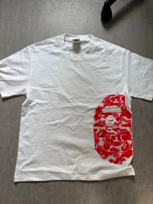 Bape t-shirt - Säljer en vit t-shirt från Bape med det klassiska ape-huvudet i röd och rosa camo på framsidan. T-shirten har rund hals, korta ärmar och är gjord i mjuk bomull. Perfekt för dig som gillar streetwear och vill sticka ut med en unik design.