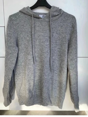 Bvaldi stickad hoodie - Mysig grå hoodie med huva och dragsnören. Tillverkad i mjukt stickat material och har en liten vit broderad logga på bröstet. Perfekt för chill dagar och enkel att matcha med jeans eller mjukisbyxor.
