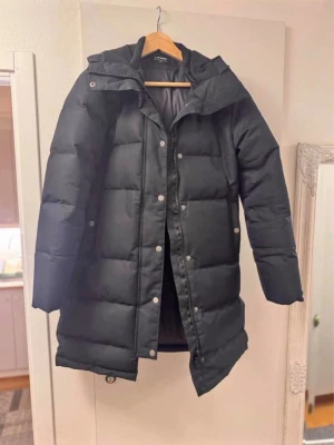 Everest vinterjacka - Säljer en svart Everest vinterjacka lång dunjacka med huva och dragkedja samt tryckknappar framtill. Jackan har en quiltad design med breda paneler och två sidofickor. Perfekt för kalla vinterdagar och riktigt varm. Enkel och stilren look som passar till det mesta.