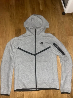 Grå Nike Tech Fleece hoodie - Snygg grå Nike Tech Fleece hoodie med hel dragkedja och svarta detaljer. Hoodien har en huva, två fickor framtill och en extra ficka med dragkedja på ärmen. Perfekt för en sportig och avslappnad stil.